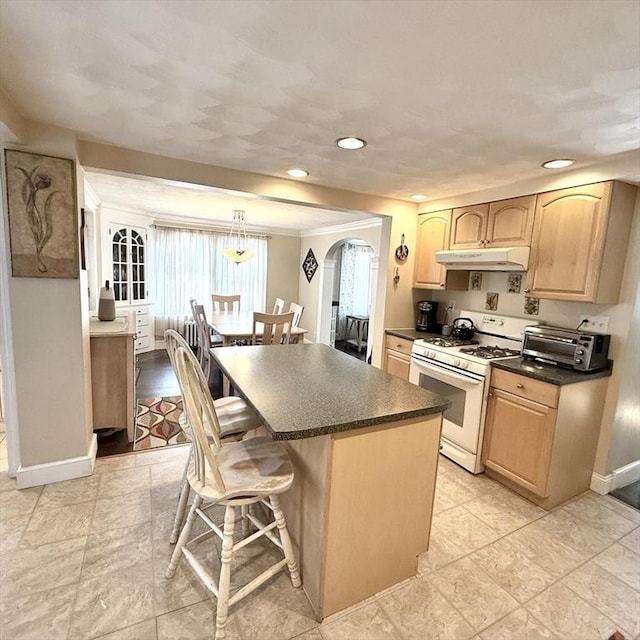 kitchen featuring white range with gas cooktop, under cabinet range hood, a breakfast bar, dark countertops, and light brown cabinets