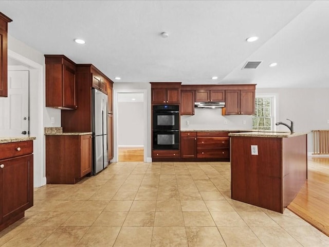 kitchen with reddish brown cabinets, recessed lighting, dobule oven black, stainless steel fridge, and under cabinet range hood