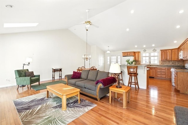 living room with baseboards, high vaulted ceiling, a skylight, ceiling fan with notable chandelier, and light wood-type flooring