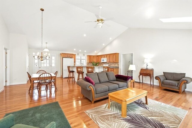 living area with baseboards, ceiling fan with notable chandelier, a skylight, light wood-style floors, and high vaulted ceiling