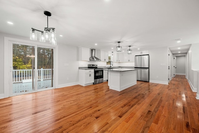 kitchen featuring wall chimney range hood, white cabinets, stainless steel appliances, decorative light fixtures, and a center island