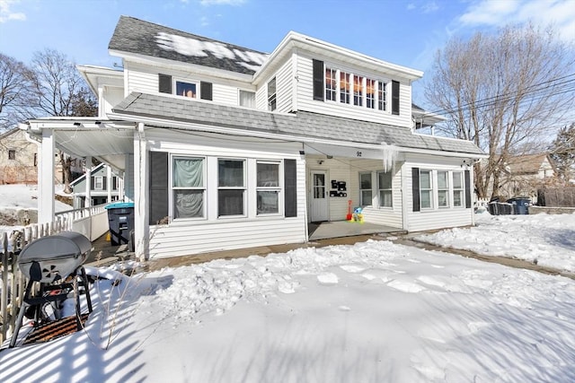 snow covered rear of property featuring covered porch, roof with shingles, and fence