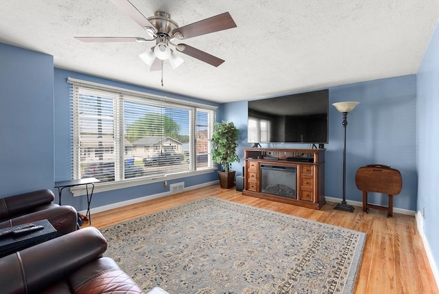 living area featuring a textured ceiling, a glass covered fireplace, ceiling fan, baseboards, and wood finished floors