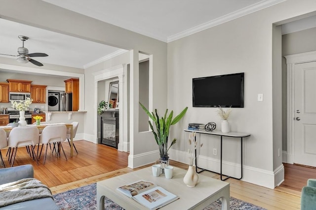 living room with crown molding, stacked washer / drying machine, ceiling fan, light wood-style flooring, and baseboards