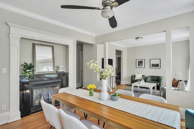 dining area featuring crown molding, a glass covered fireplace, baseboards, light wood-type flooring, and a ceiling fan