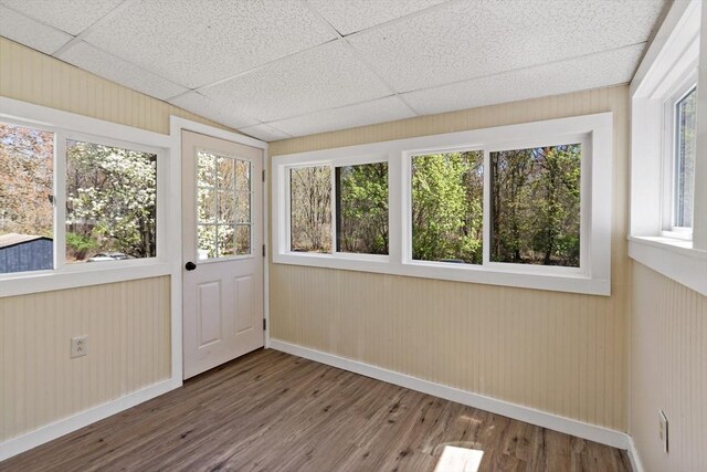 unfurnished sunroom with baseboards, a paneled ceiling, wood finished floors, and a healthy amount of sunlight