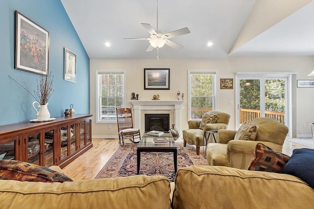 living area with baseboards, a ceiling fan, a glass covered fireplace, lofted ceiling, and light wood-style flooring