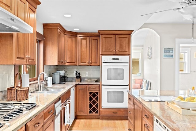 kitchen with white double oven, light stone counters, under cabinet range hood, stainless steel gas cooktop, and a sink