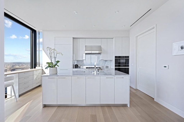 kitchen featuring tasteful backsplash, wall chimney range hood, light wood-type flooring, an island with sink, and white cabinets