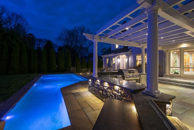 pool at night with exterior kitchen, a patio area, grilling area, and a pergola