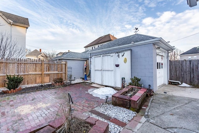 rear view of house with a patio, an outdoor structure, concrete block siding, fence, and roof with shingles