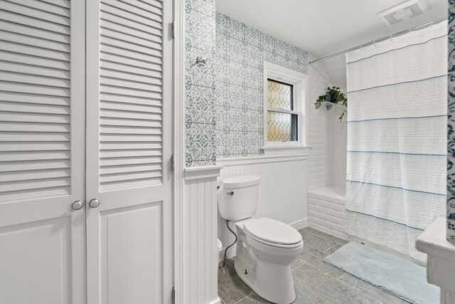 full bath with a wainscoted wall, wallpapered walls, toilet, and shower / tub combo