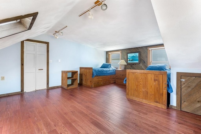 bedroom featuring rail lighting, wood finished floors, wood walls, baseboards, and vaulted ceiling