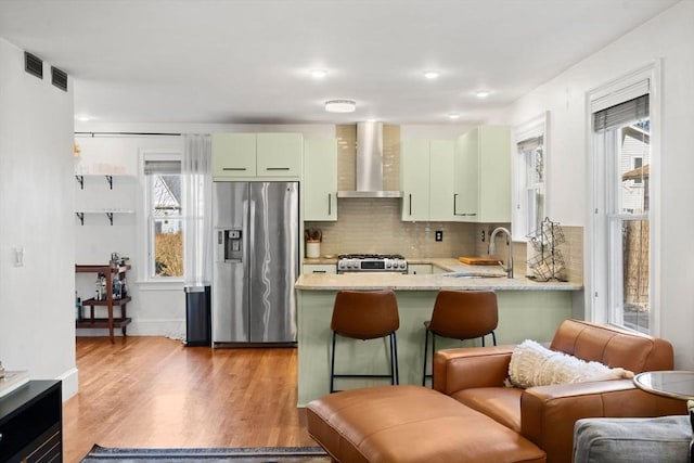 kitchen featuring a peninsula, a sink, stove, stainless steel refrigerator with ice dispenser, and wall chimney range hood