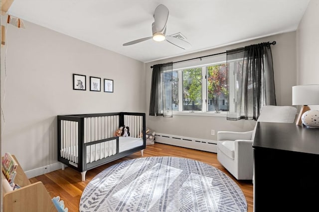 bedroom with visible vents, wood finished floors, a baseboard heating unit, a nursery area, and a ceiling fan