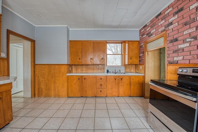 kitchen with tile countertops, a wainscoted wall, a sink, brown cabinetry, and stainless steel range with electric stovetop