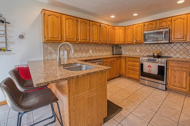 kitchen with brown cabinetry, a peninsula, light stone counters, a sink, and stainless steel appliances