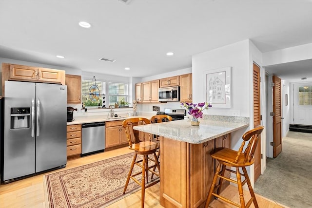 kitchen with light stone countertops, appliances with stainless steel finishes, light brown cabinets, kitchen peninsula, and a breakfast bar