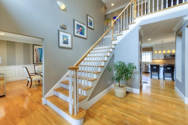 stairs with recessed lighting, a wainscoted wall, wood finished floors, a towering ceiling, and ornamental molding