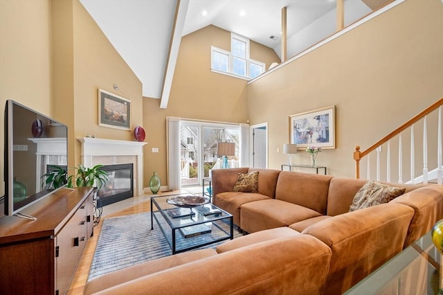 living room featuring light wood-style flooring, high vaulted ceiling, a glass covered fireplace, and stairs