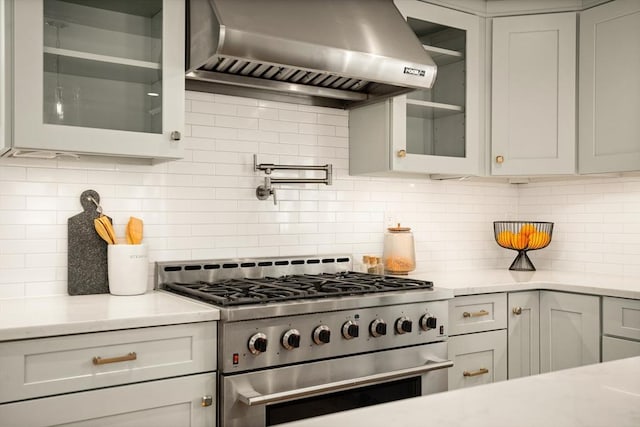 kitchen with light stone counters, decorative backsplash, wall chimney exhaust hood, stainless steel range, and glass insert cabinets