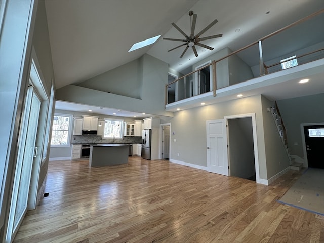 unfurnished living room with high vaulted ceiling, sink, a skylight, ceiling fan, and light wood-type flooring