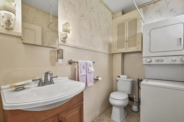 bathroom featuring wallpapered walls, vanity, tile walls, toilet, and stacked washer and clothes dryer