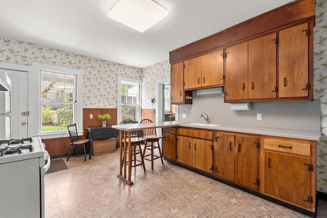 kitchen featuring wallpapered walls, light countertops, white range with gas cooktop, brown cabinets, and a sink