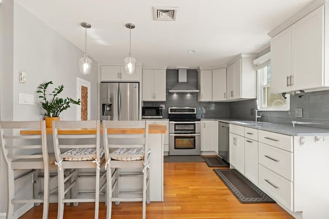 kitchen with white cabinetry, decorative light fixtures, wall chimney range hood, stainless steel appliances, and backsplash