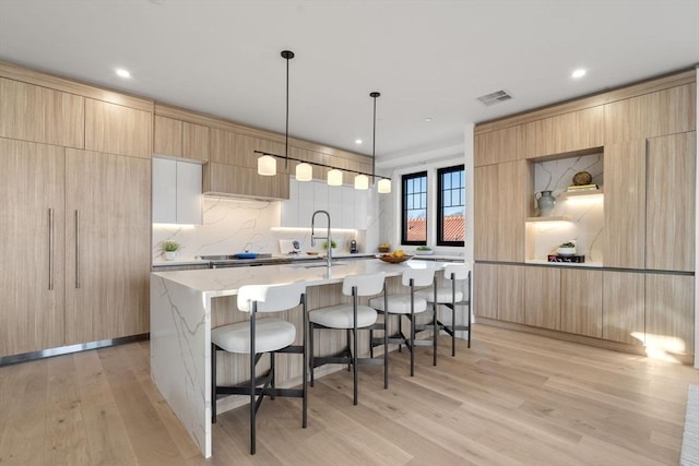 kitchen featuring light hardwood / wood-style flooring, backsplash, an island with sink, decorative light fixtures, and light brown cabinets