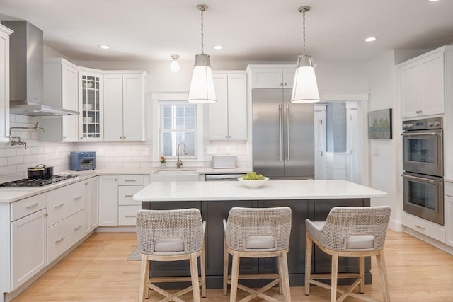 kitchen with a sink, white cabinets, wall chimney range hood, appliances with stainless steel finishes, and light wood-type flooring