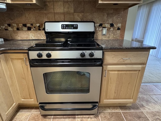 kitchen with light brown cabinets, gas stove, dark stone countertops, and backsplash