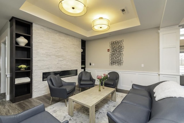 living area featuring decorative columns, a stone fireplace, wainscoting, wood finished floors, and a raised ceiling