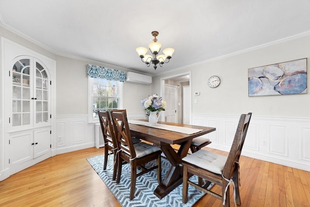 dining space featuring a wall unit AC, a notable chandelier, ornamental molding, wainscoting, and light wood-type flooring