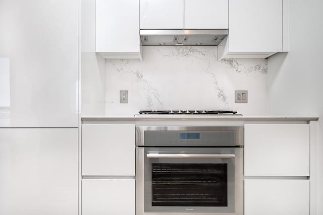 interior details featuring stainless steel oven, decorative backsplash, range hood, and white cabinetry