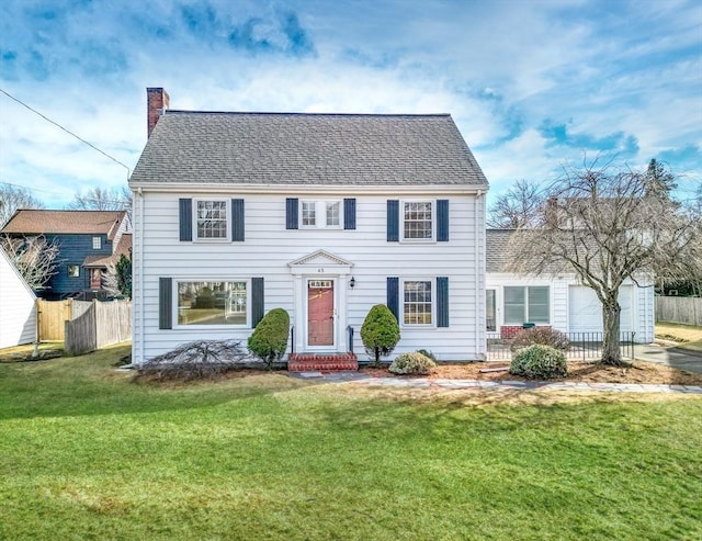 colonial home with a front yard, fence, roof with shingles, an attached garage, and a chimney