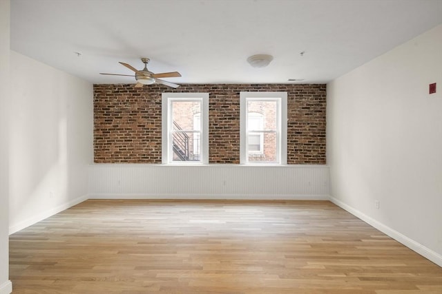 empty room with light wood-type flooring, baseboards, ceiling fan, and brick wall
