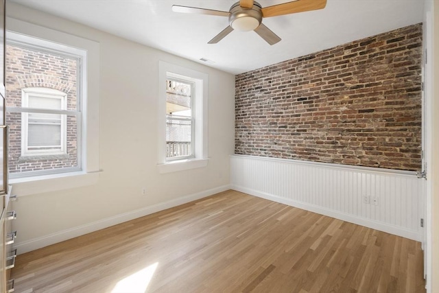 spare room with a ceiling fan, brick wall, visible vents, wainscoting, and light wood-style floors