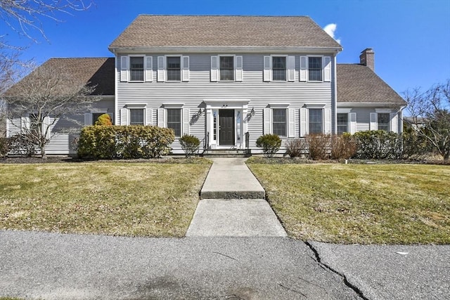 colonial home with a shingled roof, a front lawn, and a chimney