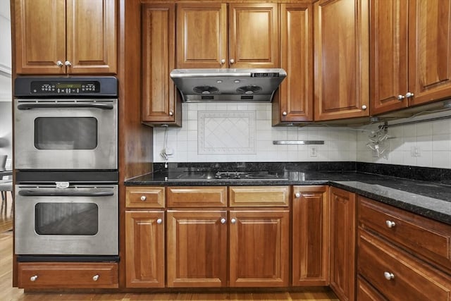 kitchen featuring under cabinet range hood, brown cabinets, double oven, and dark stone counters