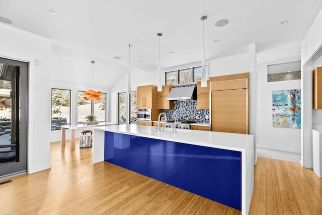 kitchen with decorative light fixtures, a kitchen island with sink, decorative backsplash, and wall chimney range hood