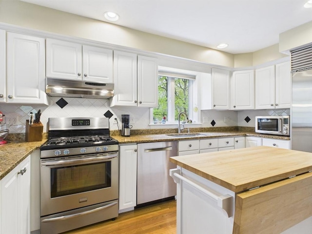 kitchen with appliances with stainless steel finishes, under cabinet range hood, butcher block counters, a sink, and white cabinetry