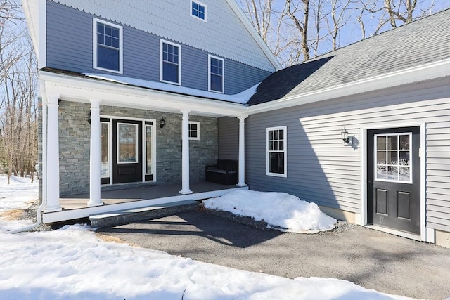 snow covered property entrance featuring covered porch, stone siding, and roof with shingles