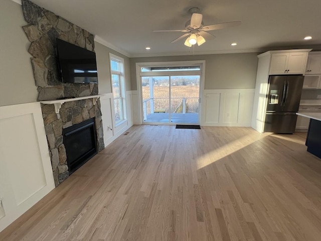 unfurnished living room with light wood-type flooring, a stone fireplace, ceiling fan, and crown molding