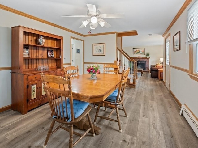 dining room featuring crown molding, light wood-style floors, a brick fireplace, a ceiling fan, and a baseboard radiator