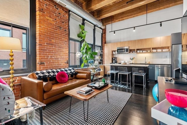 living area with beam ceiling, track lighting, brick wall, a towering ceiling, and dark wood-style flooring