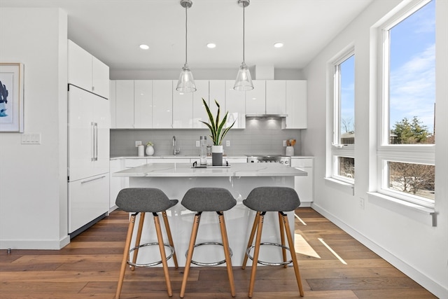 kitchen featuring paneled fridge, white cabinets, a center island with sink, and decorative backsplash