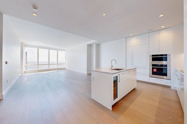 kitchen featuring stainless steel double oven, white cabinets, sink, a kitchen island with sink, and light wood-type flooring