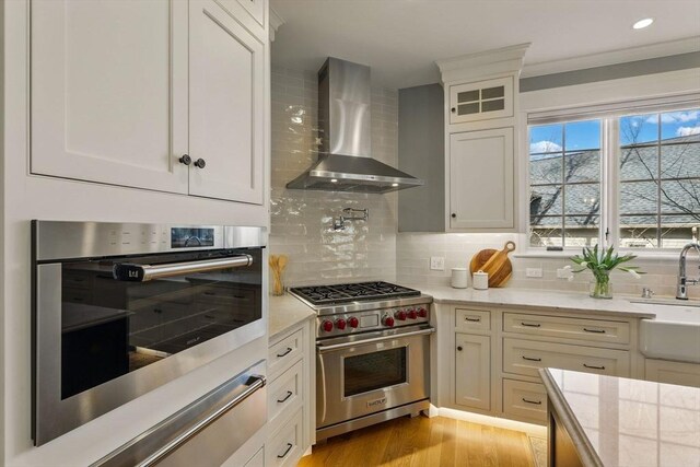 kitchen with stainless steel appliances, white cabinets, wall chimney exhaust hood, light wood-type flooring, and backsplash
