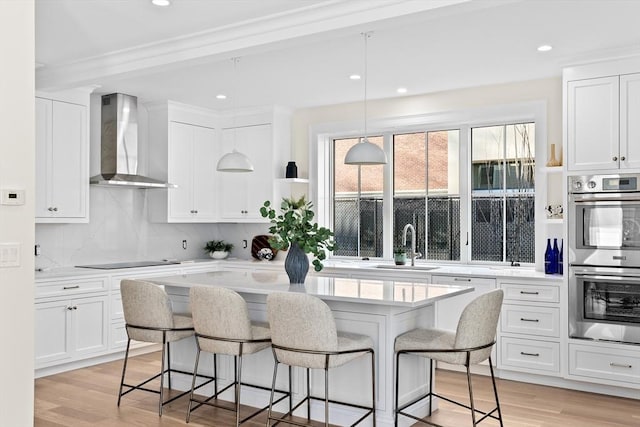 kitchen featuring black electric stovetop, wall chimney exhaust hood, light countertops, stainless steel double oven, and a kitchen breakfast bar